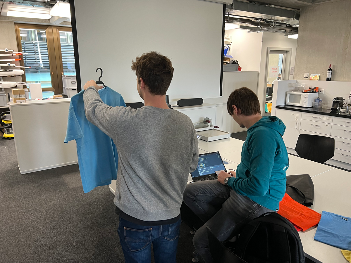 The second picture portrays a hands-on testing session in progress. In the picture, supervisor Patrick Pfreundschuh is holding a t-shirt in front of him to test the color recognition capability of Alexandru's algorithm. On the side, Alexandru is seen carefully monitoring the processing and results on his laptop, ensuring that the system accurately recognizes the shirt and its color.